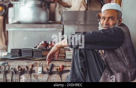 Povero vecchio pakistano triste calzolaio pakistano per le strade locali del Pakistan con le sue scarpe in pelle fatte a mano e gli attrezzi di riparazione nel suo negozio di strada Foto Stock