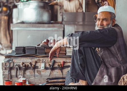 Povero vecchio pakistano triste calzolaio pakistano per le strade locali del Pakistan con le sue scarpe in pelle fatte a mano e gli attrezzi di riparazione nel suo negozio di strada Foto Stock