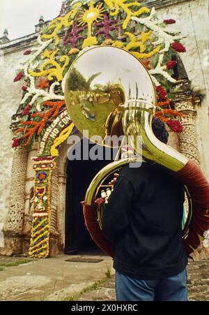 "Un uomo con un sousafone sta con le spalle allo spettatore davanti ad un cancello decorato con fiori a Ocotepec (Messico), che è stato decorato per il giorno del santo patrono della città di Ocotepec ''El Divino Salvador'' il 6 agosto 1975. [traduzione automatizzata]' Foto Stock