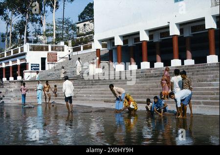 Molte persone si puliscono nel Gange lavandosi sulle rive di un fiume in una città in India. La banca è fortificata e può essere raggiunta tramite scale. Sulla riva ci sono edifici bianchi con colonne rosse. Le donne, gli uomini e i bambini stanno o squat nell'acqua poco profonda. Le donne indossano abiti leggeri colorati. [traduzione automatica] Foto Stock