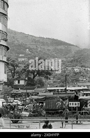 Diversi edifici di appartamenti su una strada in Hong Kong. Al centro si possono vedere capanne in legno e pietra. Tra le capanne c'è un grande albero. A sinistra di essa una parte della facciata di un grattacielo moderno. Sullo sfondo di una collina boscosa ulteriori case. [traduzione automatica] Foto Stock