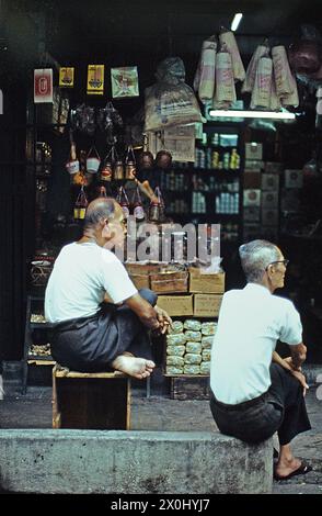 Due anziani in camicie chiare e pantaloni scuri sono seduti sul marciapiede di fronte ad un negozio a Singapore. L'uomo a sinistra nella foto è seduto su una scatola di legno o qualche tipo di sgabello, l'uomo a destra nella foto è seduto su una sporgenza di cemento. Sullo sfondo il negozio è visibile, numerosi beni appendono dal soffitto, giacciono in scatole o in piedi su scaffali. [traduzione automatica] Foto Stock