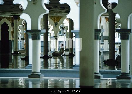 Vista tra le colonne bianche di un uomo seduto a gambe incrociate sul pavimento di una moschea a Kuala Lumpur. Il soffitto dell'edificio si riflette nel pavimento. [traduzione automatica] Foto Stock