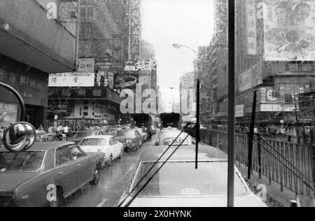 Vista lungo una strada a Hong Kong sotto la pioggia. La strada è molto trafficata e ci sono grattacieli a sinistra e a destra. I grattacieli sono coperti da grandi poster pubblicitari. La foto è stata scattata attraverso il parabrezza di un autobus. È possibile vedere uno specchietto retrovisore e i tergicristalli. [traduzione automatica] Foto Stock