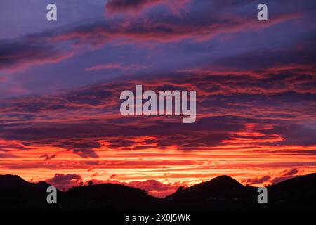 Il cielo si illumina di colori vivaci mentre il sole tramonta dietro le maestose montagne, gettando un caldo bagliore sulle soffici nuvole sparse. Foto Stock