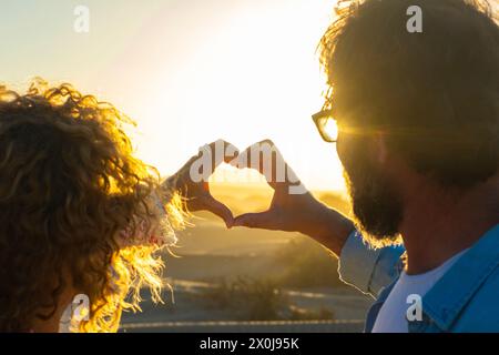Coppia romantica innamorata che fa il simbolo del cuore con le mani contro un incredibile tramonto dorato. Uomo e donna che flirtano e frequentano all'aperto godendosi la natura. La gente e la vita insieme per sempre. Giorno di viaggio Foto Stock