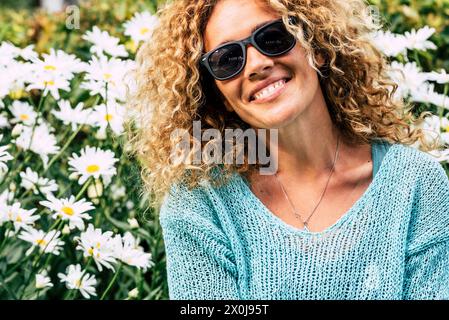 Ritratto di una bella donna adulta gioiosa con capelli ricci e occhiali da sole e margherite del parco giardino con fiori di sfondo concetto di persone e attività ricreative nella stagione primaverile. Copia spazio a sinistra Foto Stock