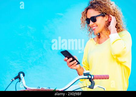 Ritratto di una bella donna adulta di mezza età sorridente al telefono cellulare seduto su una bicicletta contro una parete blu sullo sfondo. Donne e persone apprezzano la connessione tecnologica all'aperto Foto Stock