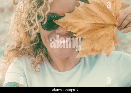 Divertente ritratto di donna felice sorridente e nascosta con una grande foglia di acero giallo asciutta autunnale. Allegri giovani adulti, donne sorridono e lok con la fotocamera indossando occhiali da sole durante le giornate di svago all'aperto Foto Stock