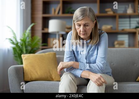 Una donna anziana siede da sola sul suo divano, guardando in basso con un'espressione triste. L'immagine cattura un momento di solitudine e di lotta emotiva spesso vissuta negli anni più anziani. Foto Stock