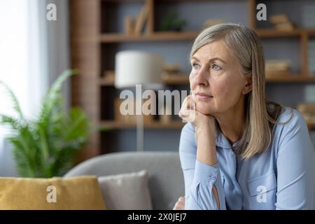 Una donna matura e serena con una camicia blu guarda con attenzione mentre è seduta in un soggiorno ben illuminato. L'atmosfera tranquilla mette in risalto il suo umore pensivo. Foto Stock