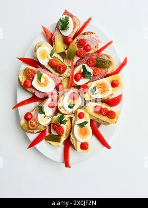 Piatto da portata con molti condimenti come pomodori, formaggio, cetriolini, fette di uova, peperoni e prosciutto cotto sul pane come antipasto Foto Stock