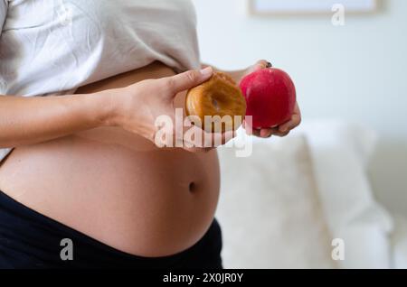 Donna incinta che esita a mangiare una ciambella o una mela. Concetto di dieta prenatale e abitudini sane. Foto Stock