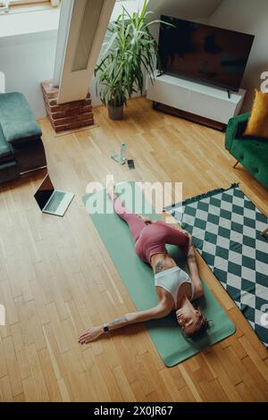 Una donna in abbigliamento sportivo si stende su un tappetino yoga sul pavimento in legno duro di un soggiorno e si allunga Foto Stock