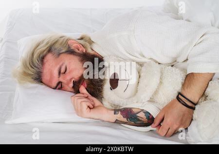 Uomo barbuto che dorme con peluche orsacchiotto che si rilassa a letto. Buona notte. Un uomo sonnolento in pigiama che dorme a letto a casa con un giocattolo per orsacchiotto. Bello Foto Stock