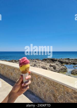 Una mano di un bambino in primo piano regge due palline di gelato in un cono, sul lungomare di Cala Ratjada, cielo blu all'orizzonte durante le vacanze estive a Maiorca, Spagna Foto Stock