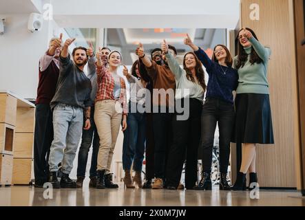 Allegro team multietnico di dipendenti che sorridono e mostrano gesti con il pollice in un luminoso spazio ufficio, illustrando il lavoro di squadra e il successo. Foto Stock
