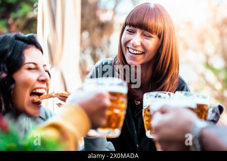 Un gruppo di amici condivide un brindisi gioioso con le tazze da birra, mentre si gusta una deliziosa pizza, - la luce del sole filtra tra tra gli alberi. Foto Stock