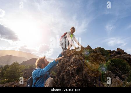 In mezzo a uno splendido paesaggio montuoso, arrampicatori maschi e femmine che raggiungono la vetta, evidenziata dalla maestosa luce del sole: Natura, avventura escursionistica e Foto Stock