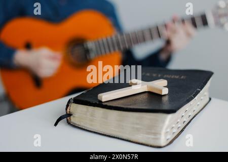 Primo piano della Bibbia cristiana su un tavolo di legno con un giovane uomo che suona la chitarra per adorare Dio in sottofondo Foto Stock