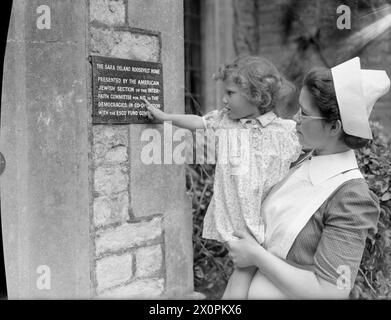 AMERICAN AID TO BRITAIN: LIFE AT SYSTON COURT NURSERY, THE SARA DELANO ROOSEVELT HOME, GLOUCESTERSHIRE, INGHILTERRA, 1942 - Un'infermiera tiene un bambino in modo che possa leggere la targa del nome della nursery in cui risiede. La targa recita: "The Sara Delano Roosevelt Home. Presentato dalla sezione ebraica americana del Comitato interreligioso per gli aiuti alle democrazie in cooperazione con il Comitato del Fondo esco" Foto Stock