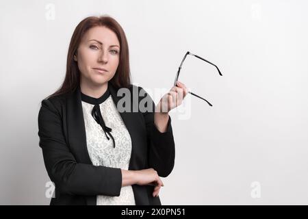 Donna d'affari sicura che tiene in mano gli occhiali e guarda la macchina fotografica. La donna caucasica Gen X è vestita con abiti da ufficio e si erge su sfondo bianco Foto Stock