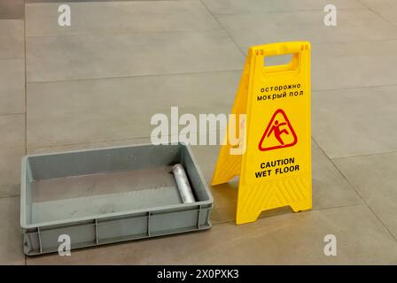 Accanto a un vassoio d'acqua in plastica grigia è presente un cartello giallo per il pavimento bagnato Un panno sporco su un pavimento piastrellato, per garantire che le misure di sicurezza siano applicate Foto Stock