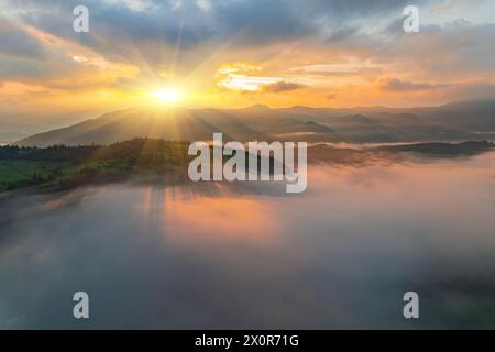 La nebbia avvolge la foresta di montagna. I raggi del sole nascente si infrangono attraverso la nebbia. Vista aerea del drone. Foto Stock