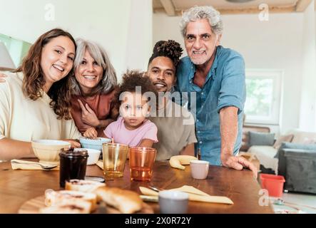 Una famiglia diversificata e multigenerazionale si riunisce intorno al tavolo della cucina, condividendo un momento di gioia: Nonni, genitori e un bambino sorridono calorosamente, reinfor Foto Stock