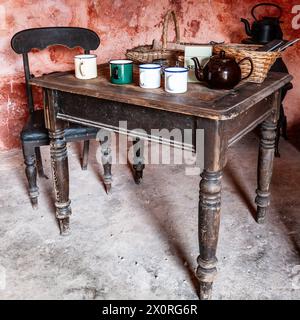 Antico tavolo in legno in un ufficio agricolo, con tazze di latta, teiera marrone e cesti di vimini. Pavimento sporco e colori delle pareti sbiaditi. Stile retrò. Foto Stock