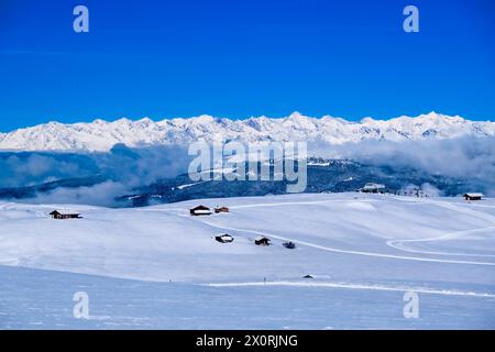 Campagna agricola collinare con pascoli innevati e capanne in legno all'Alpe di Siusi in inverno, vette della catena principale delle Alpi in distan Foto Stock