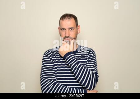 Uomo ispanico con barba di circa 40 anni che indossa un maglione a righe pensando concentrato nel dubbio con il dito sul mento e guardando la fotocamera Foto Stock