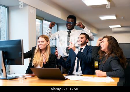 Il team di uffici multietnici si rallegra intorno al computer, il buon dirigente africano alza i pugni in vittoria, i colleghi tifanno, l'uomo punta sullo schermo mostrando la crescita Foto Stock
