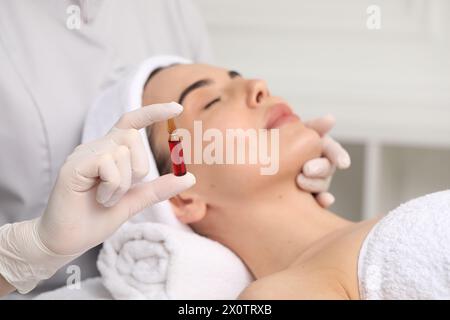 Cosmetologo professionista che tiene in mano una fiala per la cura della pelle mentre lavora con il cliente in clinica, primo piano Foto Stock