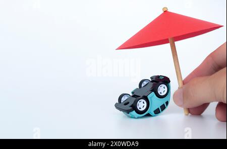 Mano che tiene l'ombrello giocattolo rosso e l'auto capovolta su sfondo bianco. Concetto di copertura assicurativa. Foto Stock
