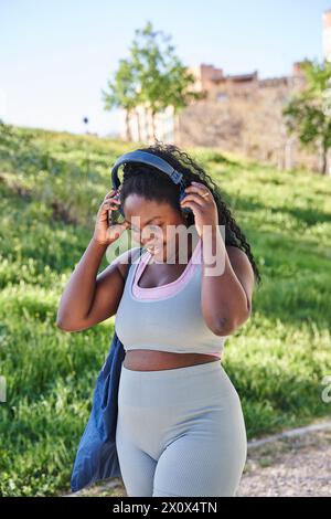 Una donna afroamericana di successo in un parco durante l'esercizio fisico attivo e il jogging utilizza l'app per smartphone, una donna sportiva che sorride con le cuffie in ascolto Foto Stock