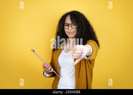 Donna d'affari afroamericana con documenti in mano su sfondo giallo con faccia arrabbiata, segno negativo che mostra antipatia con il pollice giù Foto Stock