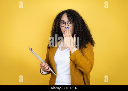Donna d'affari afroamericana con documenti in mano su sfondo giallo che copre la bocca con la mano, scioccata e spaventata per errore. sorpreso Foto Stock