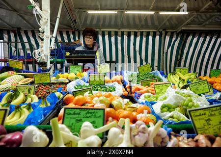 Reichhaltige Obst- und Gemüseauswahl an einem Marktstand. Eine Verkäuferin steht im Hintergrund an einer Waage und wiegt aus. Foto Stock
