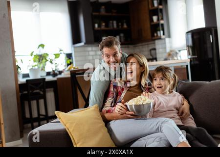 Una famiglia di tre persone si trova comodamente adagiata su un divano, con i volti che riflettono l'emozione e l'attenzione mentre condividono una ciotola di popcorn durante una sosp Foto Stock