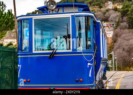 Tram blu con il numero sette ai suoi lati che viaggiano su una strada che sale su una collina, turista femminile all'interno che guarda la macchina fotografica, montagne con casa sfocata Foto Stock