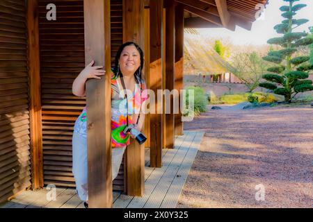 Allegra donna di mezza età che abbraccia una colonna di legno e guarda la macchina fotografica, in piedi sulla veranda della casa di campagna, la macchina fotografica appesa al collo, S. Foto Stock