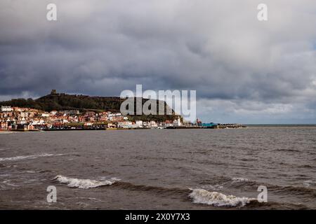 Piccole onde si infrangono sulla spiaggia con nuvole scure su una città costiera illuminata dal sole con castello su una collina, Scarborough, North Yorkshire, Inghilterra. Foto Stock