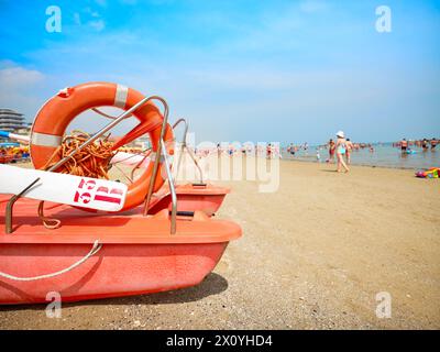 Scialuppa di salvataggio in primo piano sulla spiaggia in una soleggiata mattinata d'estate, sullo sfondo alcuni bagnanti si rinfrescano sulla costa adriatica, in Italia. Foto Stock