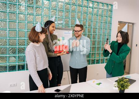 Allegra e attraente donna afroamericana che apre un regalo dato dai colleghi multietnici in carica per applaudire i colleghi. Felice squadra di giovani Foto Stock