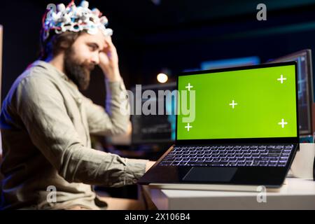 Concentratevi sul laptop con schermo verde utilizzato dagli sviluppatori che indossano un auricolare EEG, traducendo i pensieri in comandi del PC. Professionista IT che controlla le funzioni del computer utilizzando Mind, aiutato da un notebook mockup Foto Stock