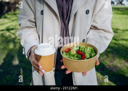 Una donna regala un caffè da asporto e una ciotola di vibrante insalata verde mista con fragole fresche, pronti per un delizioso pasto in un parco illuminato dal sole Foto Stock