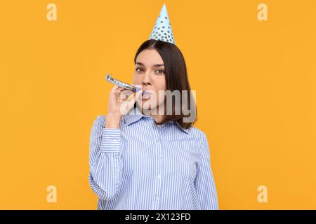 Giovane donna in cappello da festa con soffiatore su sfondo giallo Foto Stock