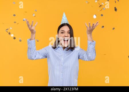 Giovane donna felice in cappello da festa vicino a coriandoli volanti su sfondo giallo Foto Stock