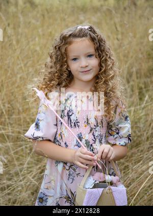 Bella ragazza dai capelli ricci vestita con un abito rosa che regge l'estate a forma di stella e una borsa in un campo coltivato con erba marrone pastello Foto Stock